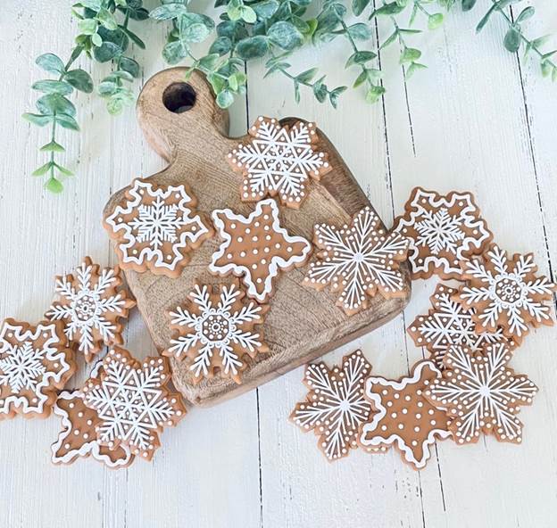 May include: A wooden cutting board with twelve gingerbread cookies shaped like snowflakes. The cookies are decorated with white icing and are arranged in a circular pattern on the board. The board is on a white wooden surface.