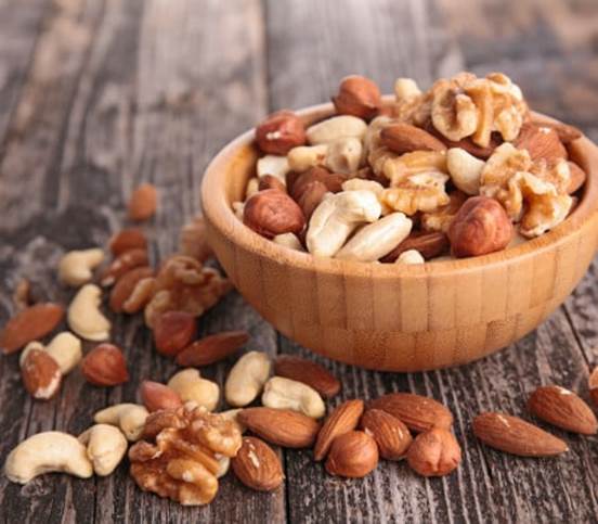 A wooden bowl filled with a variety of Raw Nuts, including almonds, cashews, walnuts, and hazelnuts, placed on a rustic wooden table with some nuts scattered around.