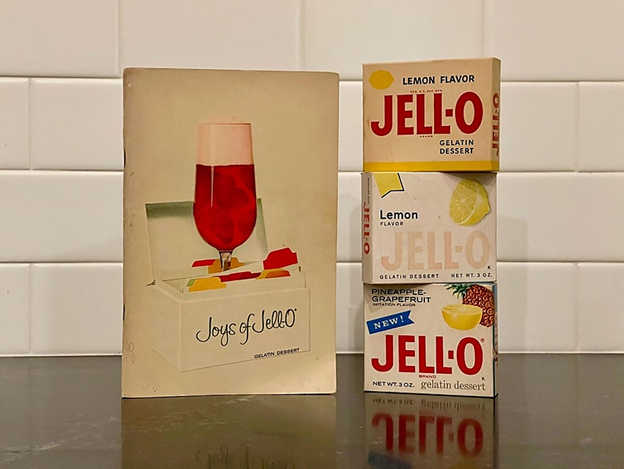 May include: A vintage Jell-O box set with three boxes stacked on top of each other. The top box is lemon flavored, the middle box is lemon flavored, and the bottom box is pineapple-grapefruit flavored. The boxes are all white with red and yellow lettering. The boxes are sitting on a brown surface.