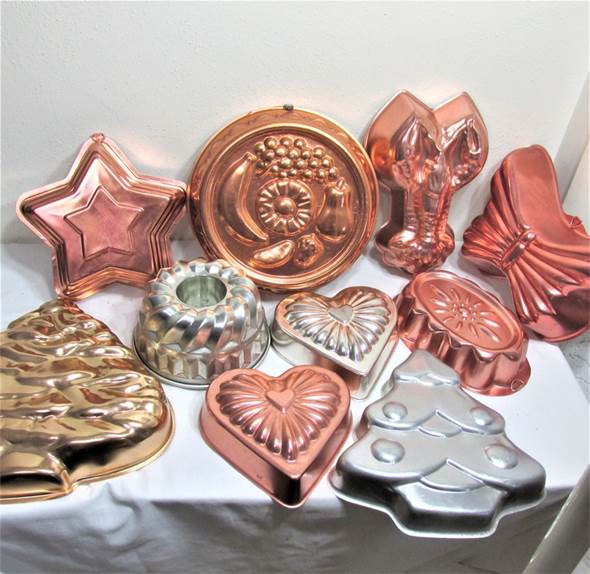 May include: A collection of various metal baking molds in copper and silver tones, featuring shapes such as a star, a round fruit platter, a lobster, a bow, a Christmas tree, a heart, and a fluted ring mold, all arranged on a white surface.